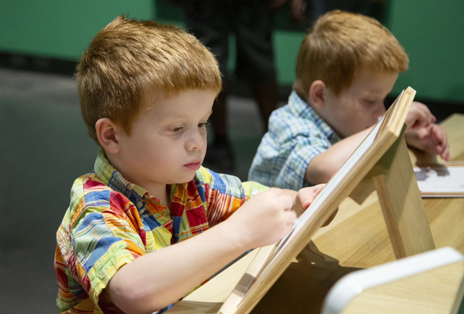 Two young boys with red hair focus on drawing at wooden easels. One wears a colorful plaid shirt, while the other is in a blue plaid shirt. They are seated next to each other in a classroom setting with green walls.