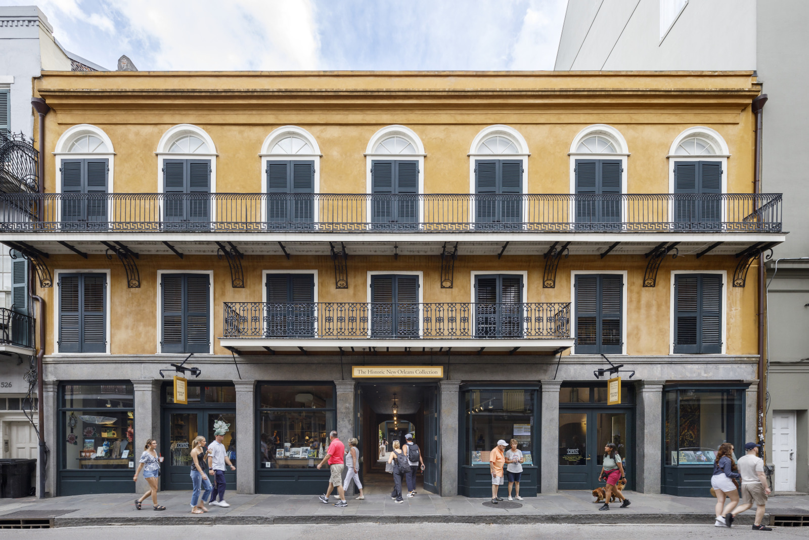 A two-story yellow building with arched windows and black shutters stands on a busy street. People walk by, with some stopping to look at the storefront displays. A black wrought-iron balcony decorates the facade above.