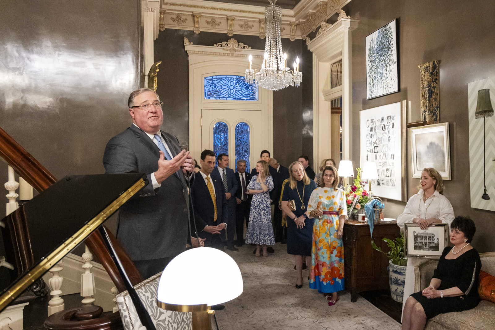 A man is speaking in a well-decorated room to a group of people. The audience is standing and listening attentively. There is a chandelier on the ceiling and art on the walls. Some attendees hold drinks, and one holds a picture frame.