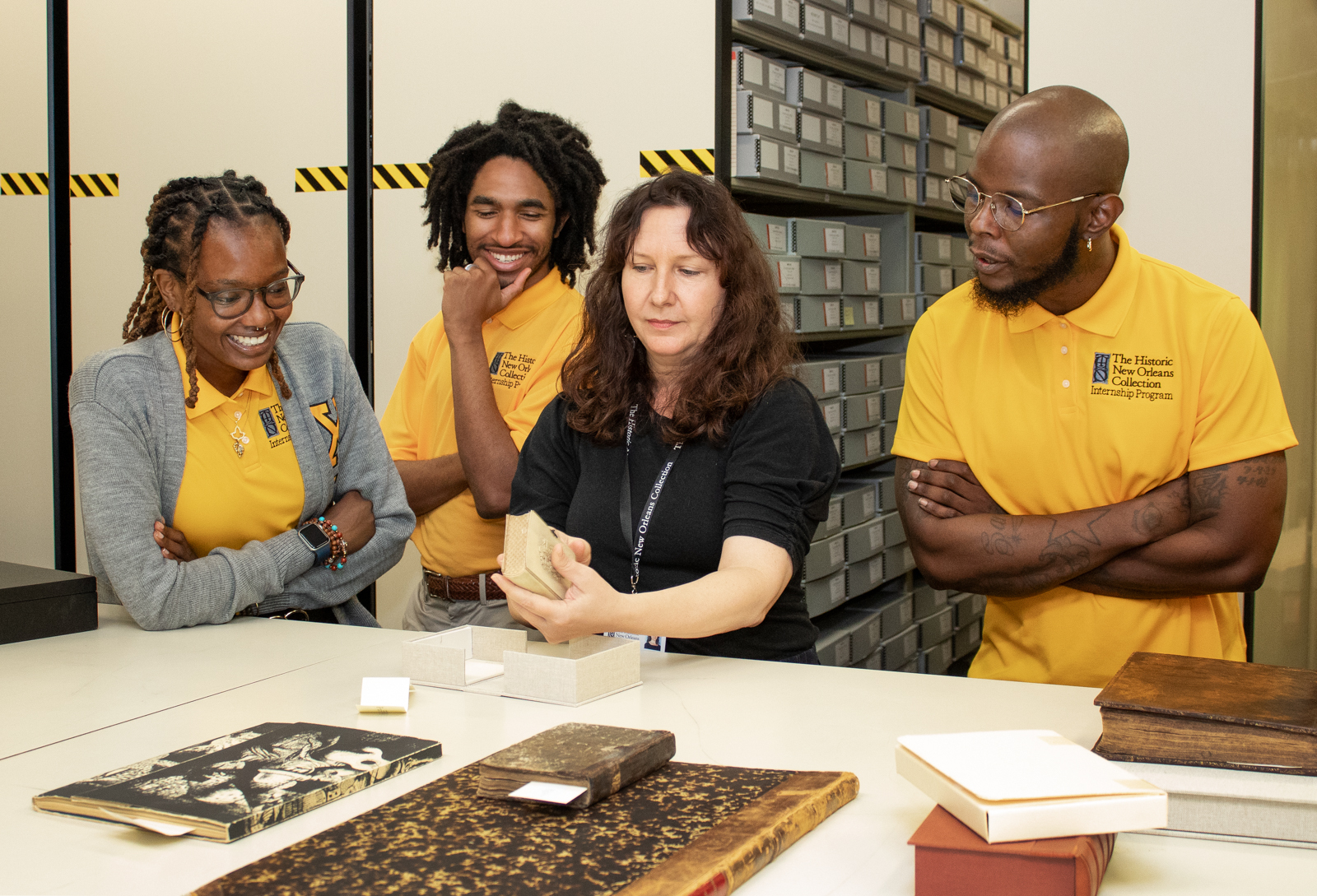 A group of three people in yellow shirts and one woman in a black shirt are gathered around a table with historical books and documents. The woman shows a book to the group in an archival setting. Shelves filled with boxes are in the background.