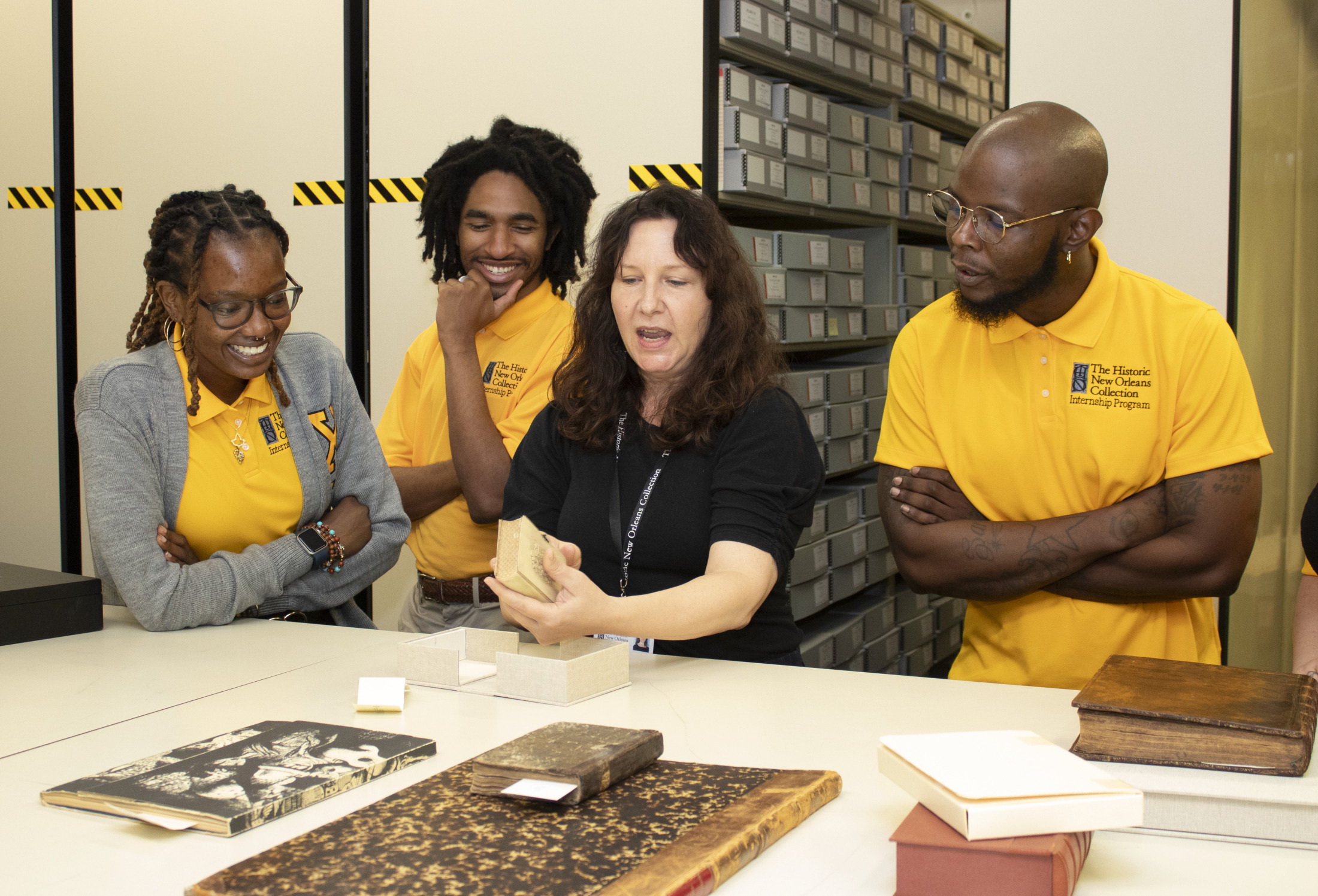 A group of four people, three wearing yellow shirts, are gathered around a table in an archives room. They are looking at and discussing a historical document shown by a person in a black shirt. Shelves filled with archival boxes are in the background.