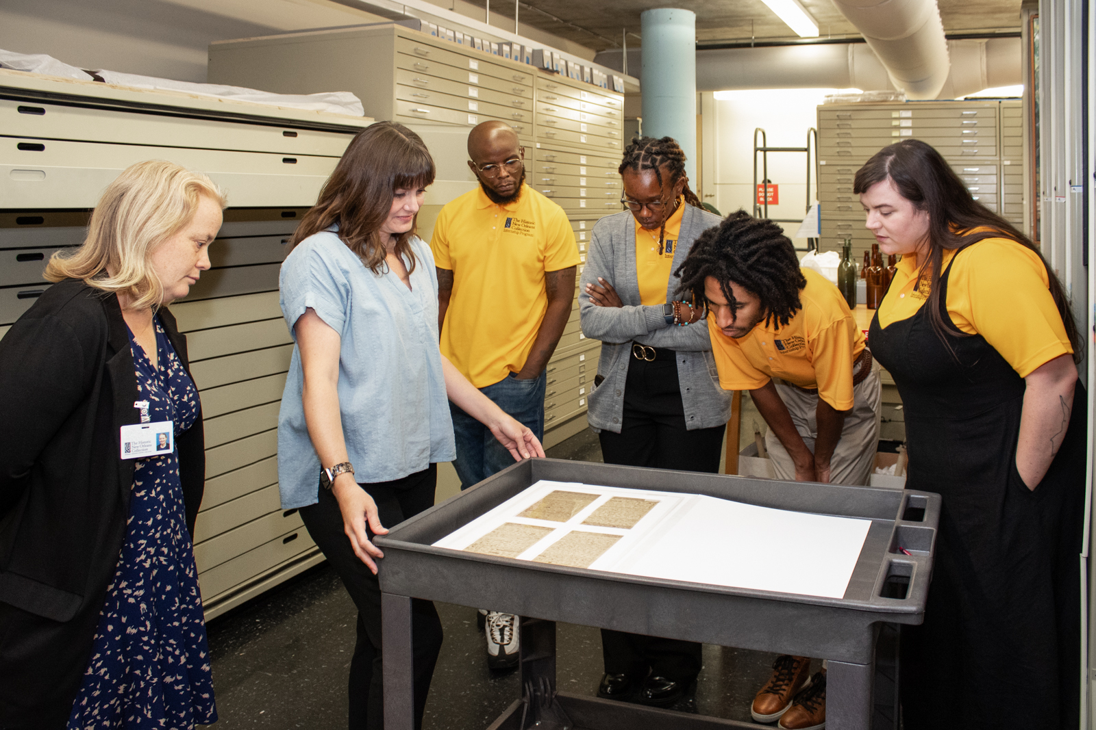 A group of six people gathers around a cart with historical documents in a storage room. Two people wear ID badges, and others wear yellow shirts with logos. They appear focused and engaged in examining the items. Shelves line the walls.