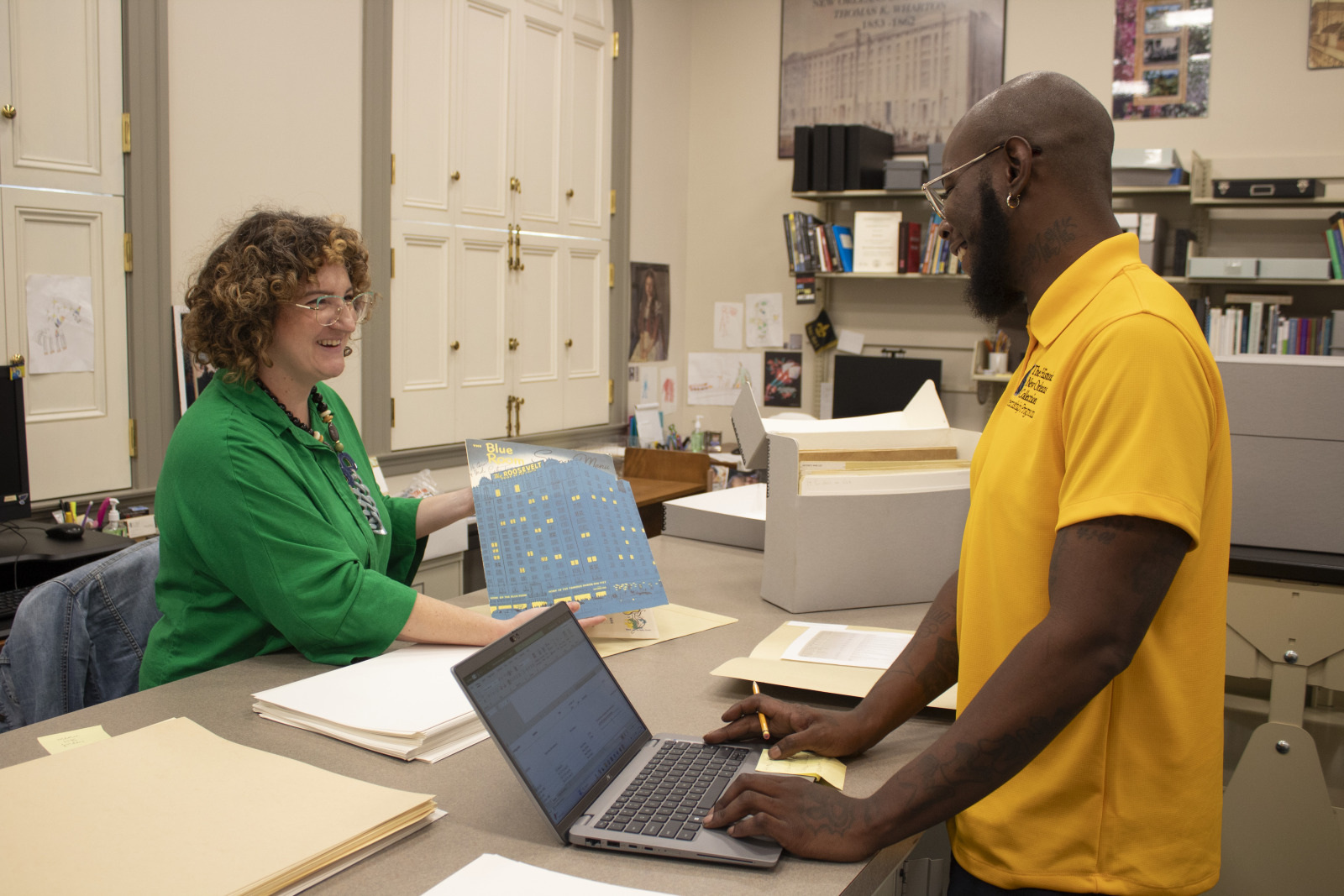 Two people in a library interact at a desk. The person on the left holds a colorful sheet, smiling, while the person on the right, in a yellow shirt, looks at a laptop. Shelves with books and documents are visible in the background.