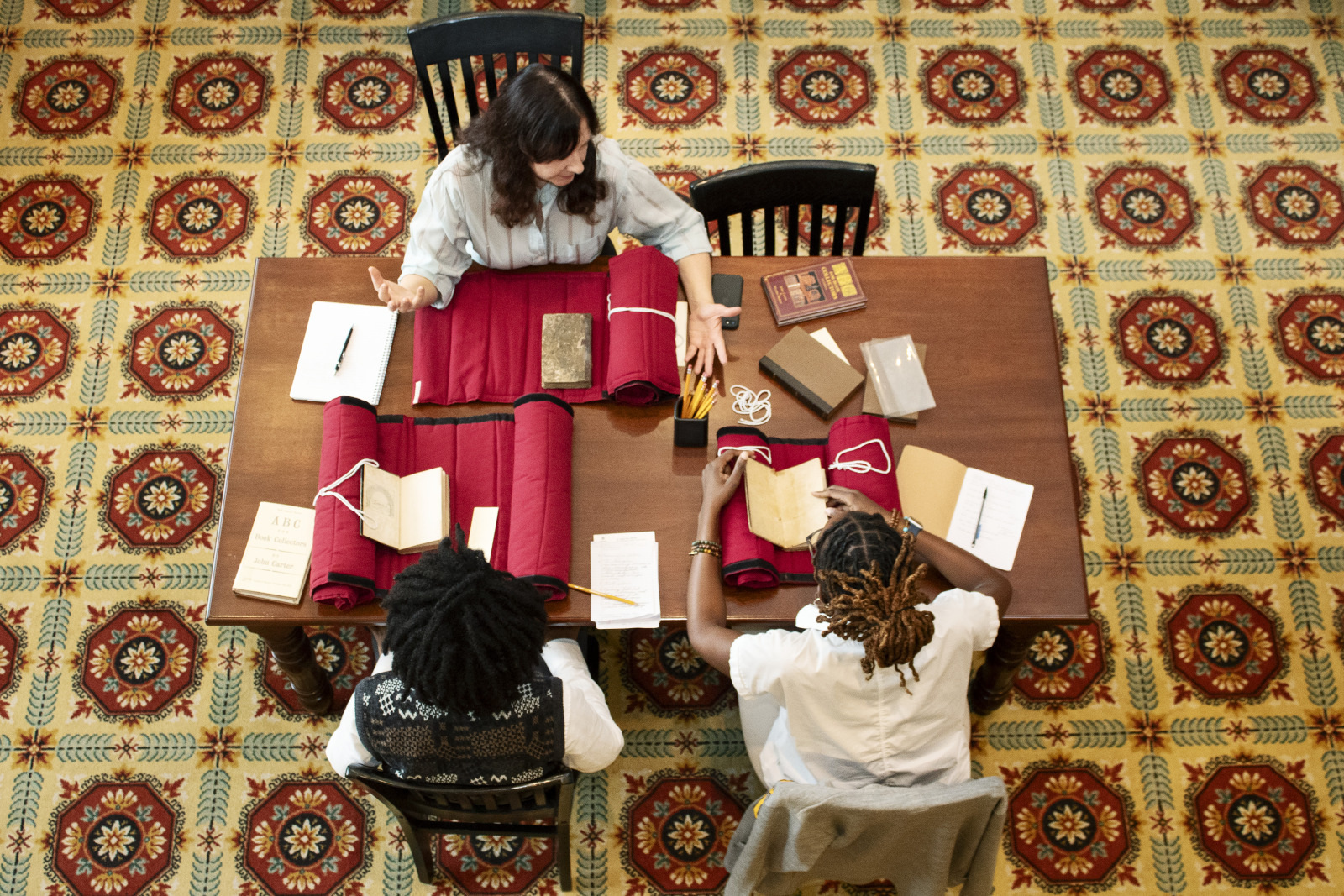 Three people sit at a wooden table with books and red folders open. They are engaged in discussion, surrounded by notebooks and other documents. The floor has an intricate, colorful patterned carpet. The scene is viewed from above.