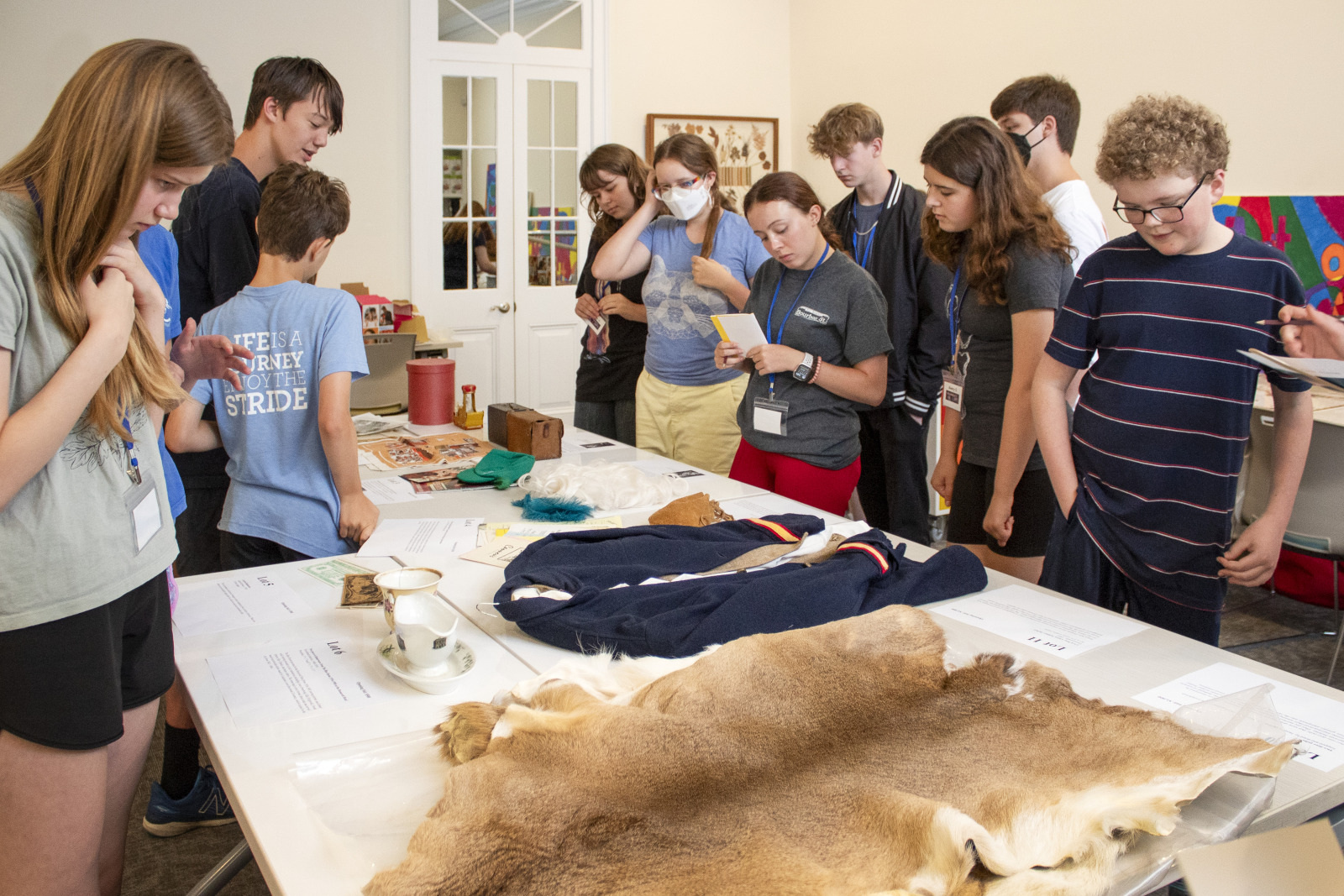 A group of students gather around a table displaying historical artifacts and animal pelts. They are reading and discussing various items, with some taking notes. The room is well-lit and educational in atmosphere.