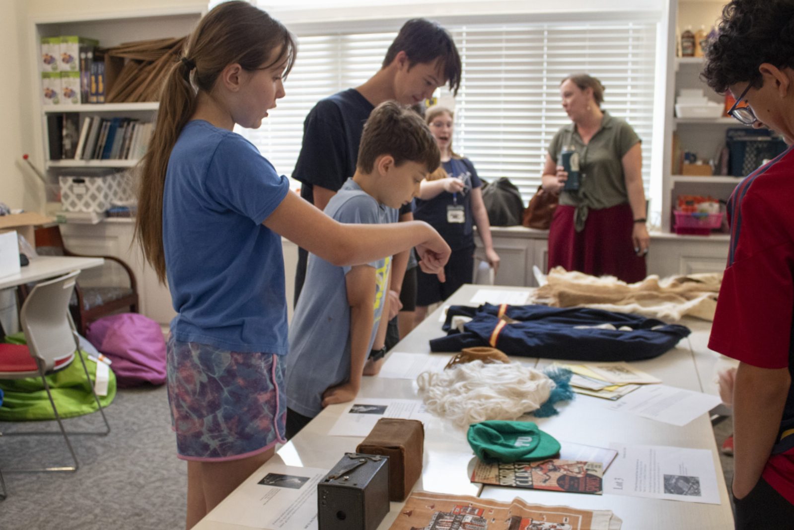 Children and adults in a classroom engage with a display table featuring various educational items. The room is bright with natural light from a window, and shelves with books and supplies are visible in the background.