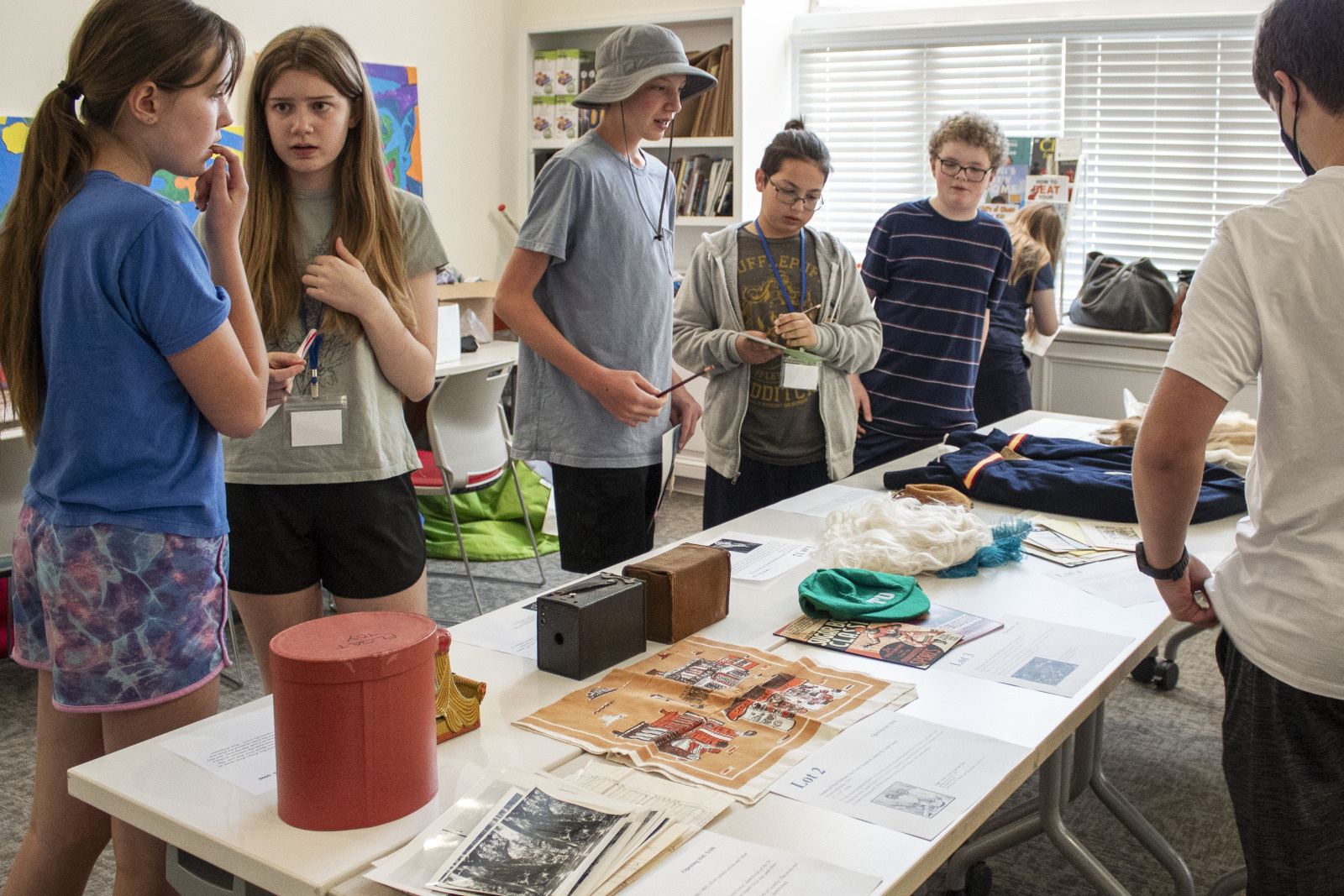 A group of children stand around a table covered with vintage items, documents, and photos. The room is well-lit with natural light from a window. The children appear engaged and are discussing the displayed objects.