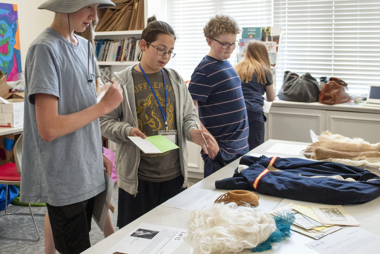 Three people examine items on a table, including costumes and documents. One person takes notes, while another gestures at the items. Shelves with books and a window are in the background.