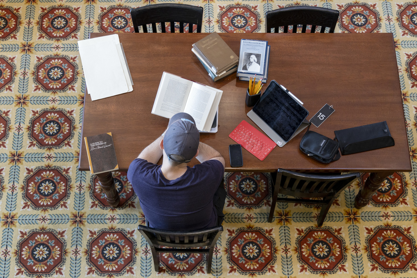 A person wearing a cap sits at a wooden table, reading a book in a library. The table is spread with more books, a tablet, a smartphone, a pencil holder, and a notebook. The floor has a patterned carpet. The date on the tablet reads 16.