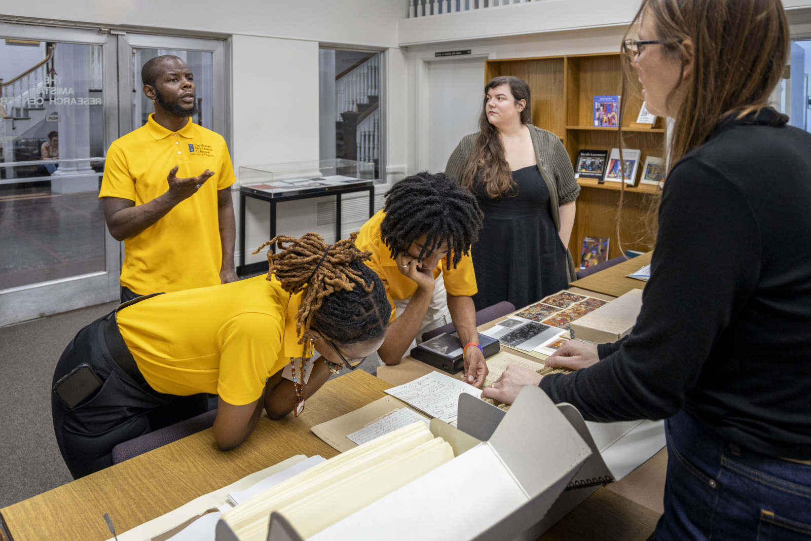 A group of people are gathered around a table in a library-like room. Three individuals in yellow shirts are examining documents, while two others, one standing and one seated, look on. Shelves with books and stairs are visible in the background.