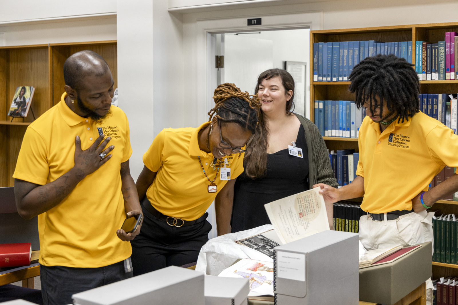 Four people, three wearing matching yellow polo shirts, are gathered around historical documents in a library. They appear engaged and interested, with shelves of books in the background.