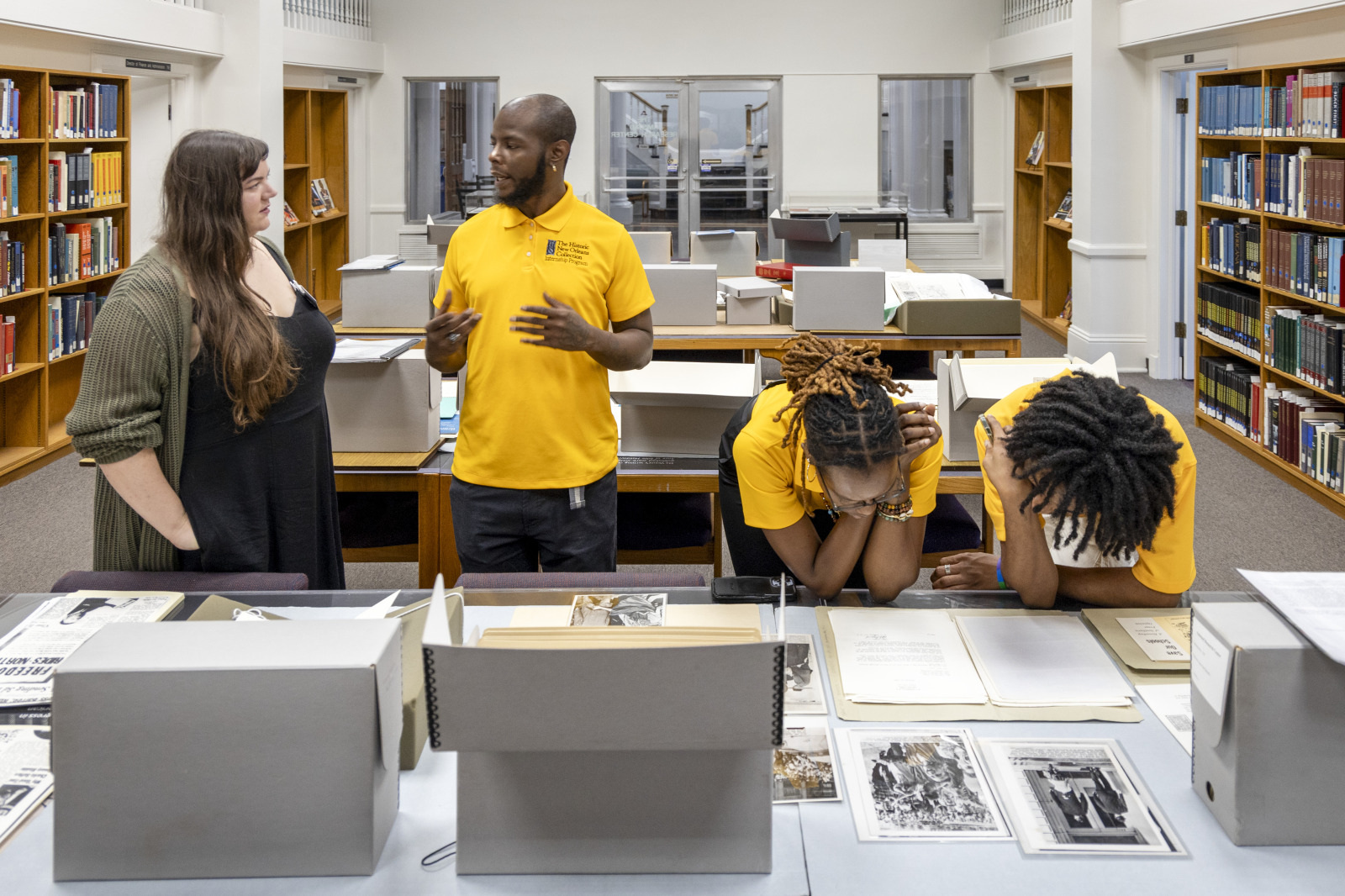 A man in a yellow shirt speaks to a woman in a library. Two other people, also in yellow shirts, lean over a table looking through documents and photographs. Bookshelves line the walls around them.