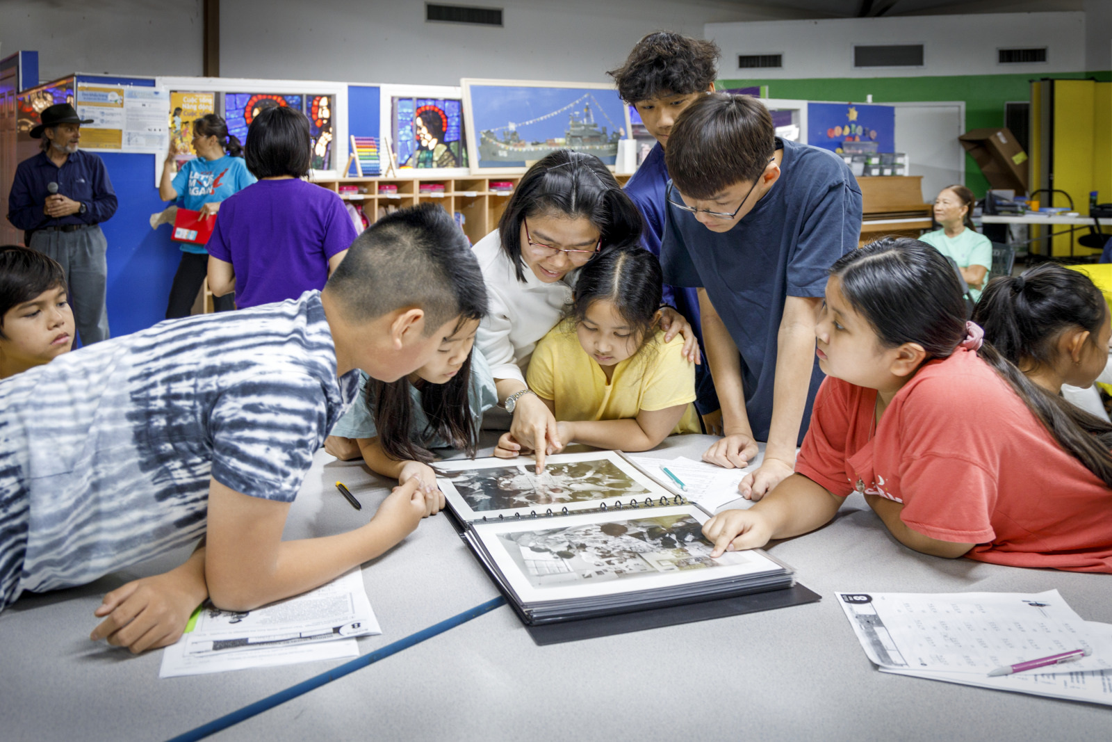 A group of children and a teacher gather around a table, looking at a photo album. The classroom setting is colorful, with educational posters on the walls. The children appear engaged and curious.