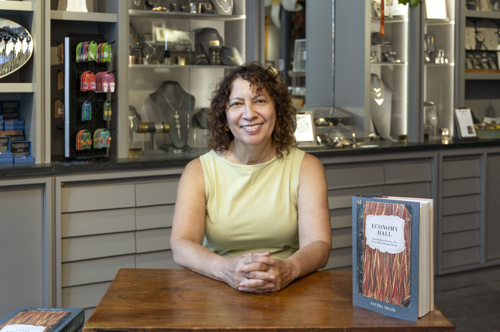 A woman with curly hair sits at a wooden table, smiling. She wears a light yellow sleeveless top. In front of her is a book titled Economy Hall. The background shows shelves filled with various items.