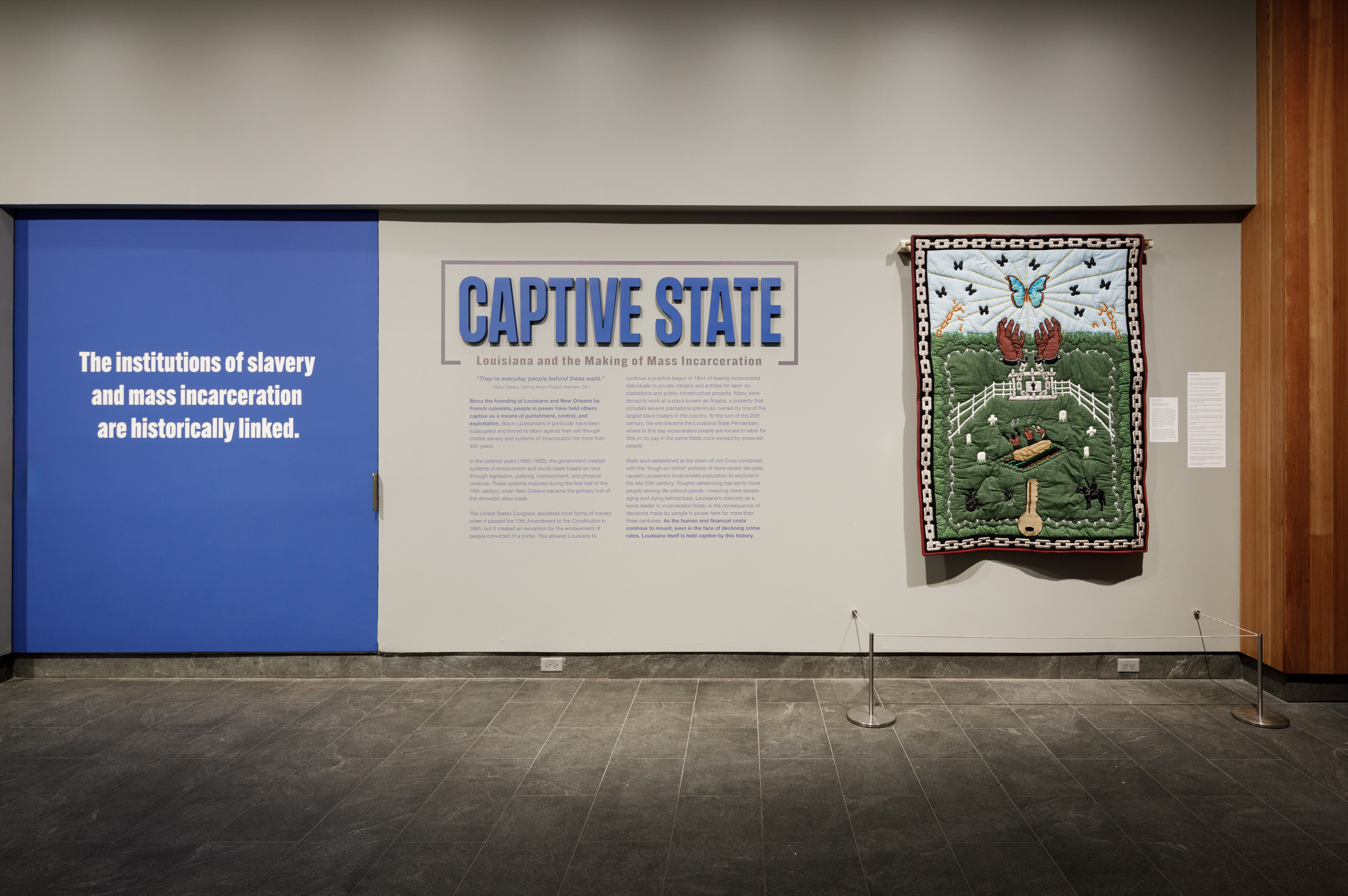 Exhibit wall with a blue panel stating, The institutions of slavery and mass incarceration are historically linked. A section titled CAPTIVE STATE features text and a colorful quilt with nature and wildlife motifs displayed on the right.