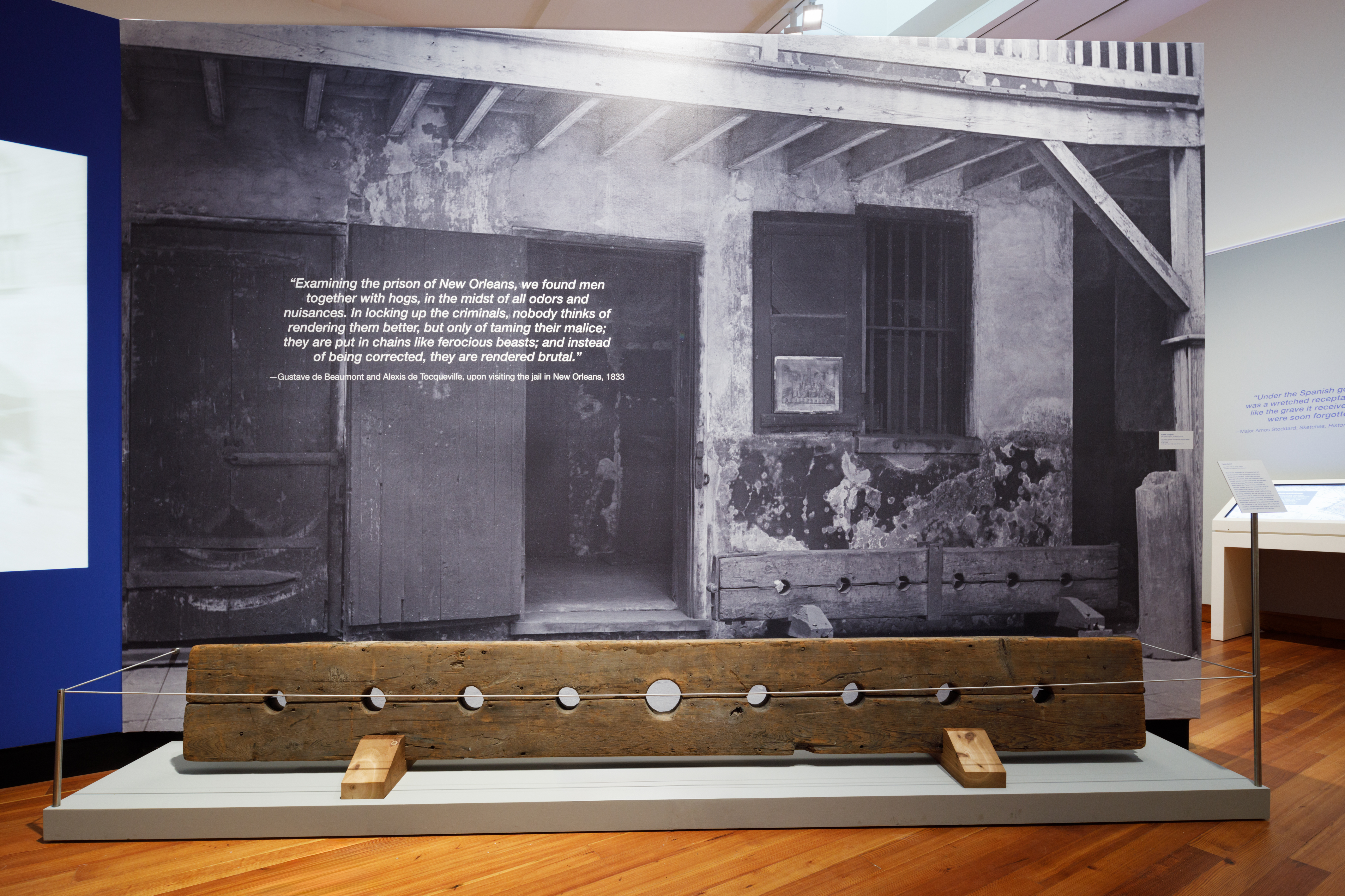 A historical museum exhibit features a large black-and-white photograph of an old prison building. In front, there is a wooden stock used for restraining prisoners, with openings for neck and wrists. The quote on the photo describes the harsh prison conditions.