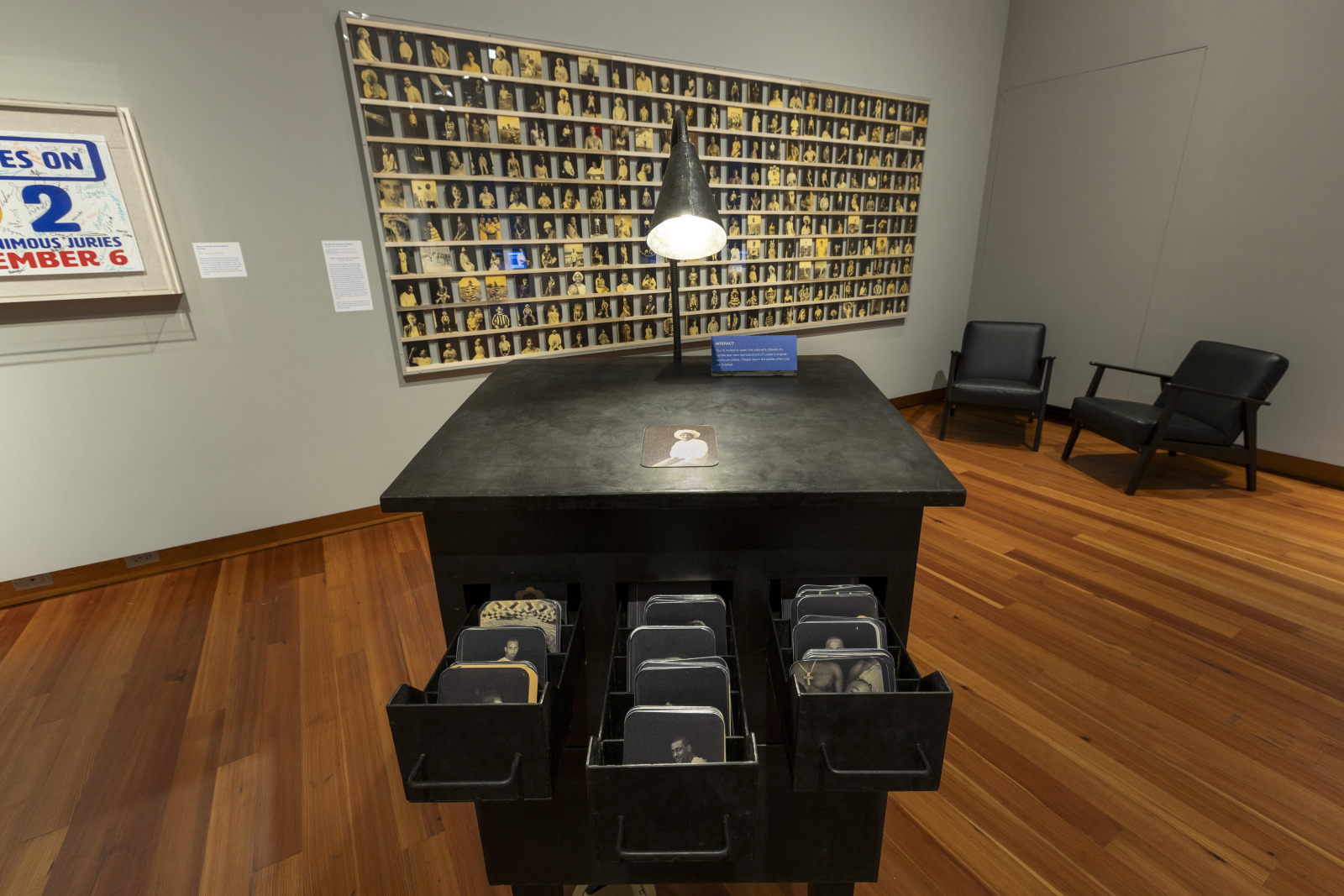 A photograph shows a metal cabinet with draws full of photographs in the middle of an exhibition gallery floor, with a large portrait collage showing images of incarcerated people mounted on a wall behind it.