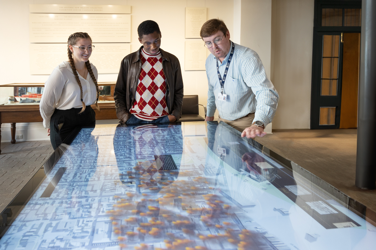 Three people stand around an interactive digital table displaying a city map. One person gestures to the map, engaging the others, who look on with interest. The setting appears to be an educational or museum space.