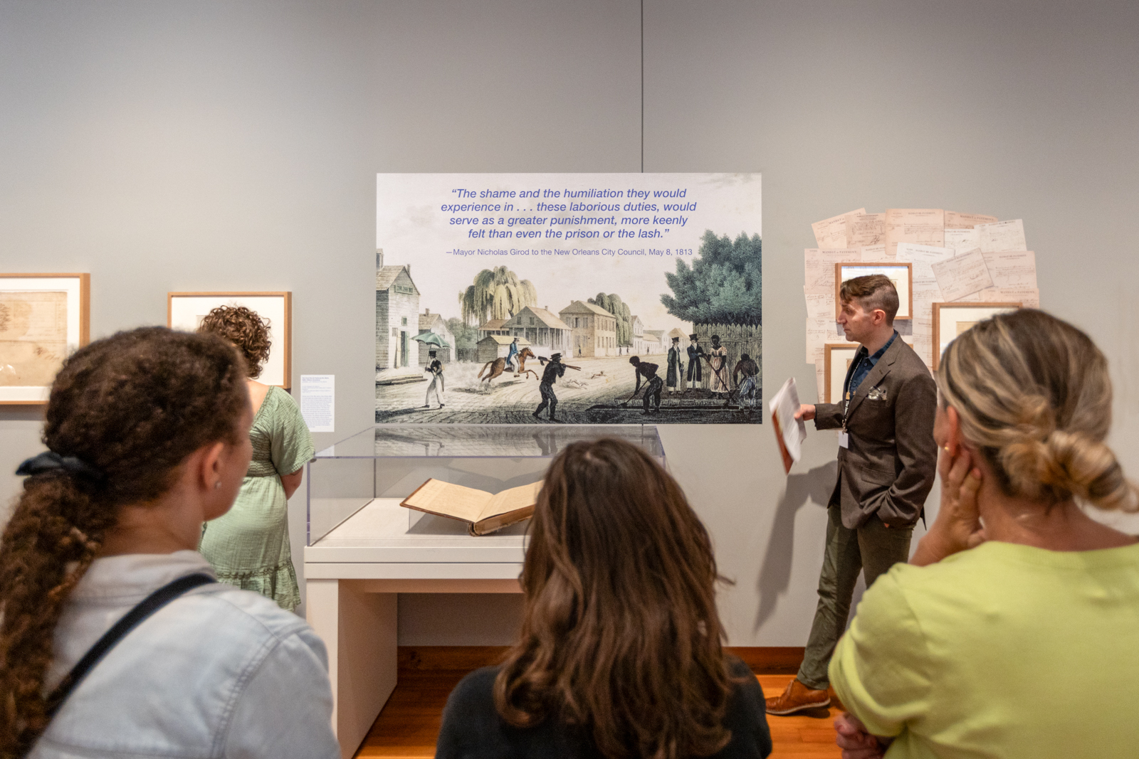 A group of people view an exhibit in a gallery featuring a large historical illustration and several framed documents. A guide discusses the display, which includes a quote about shame and humiliation in a prison setting.