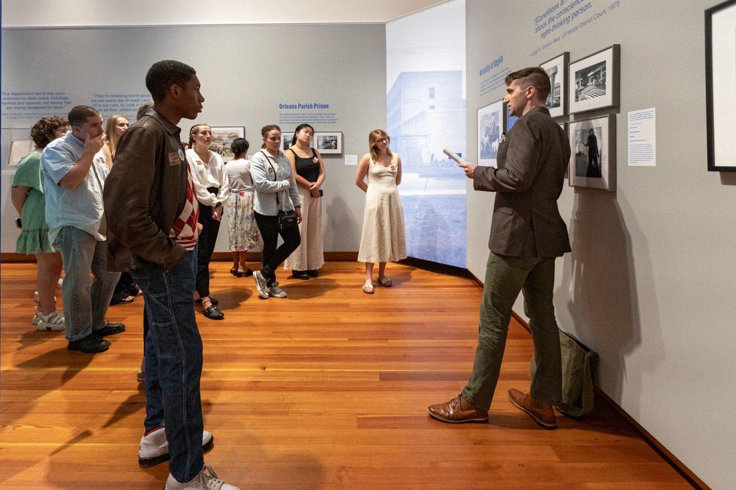 A group of people in a gallery listens to a man standing near framed photographs on a wall. The man appears to be explaining the artwork. The floor is wooden, and the walls are gray with blue text. Some people are taking notes or photos.