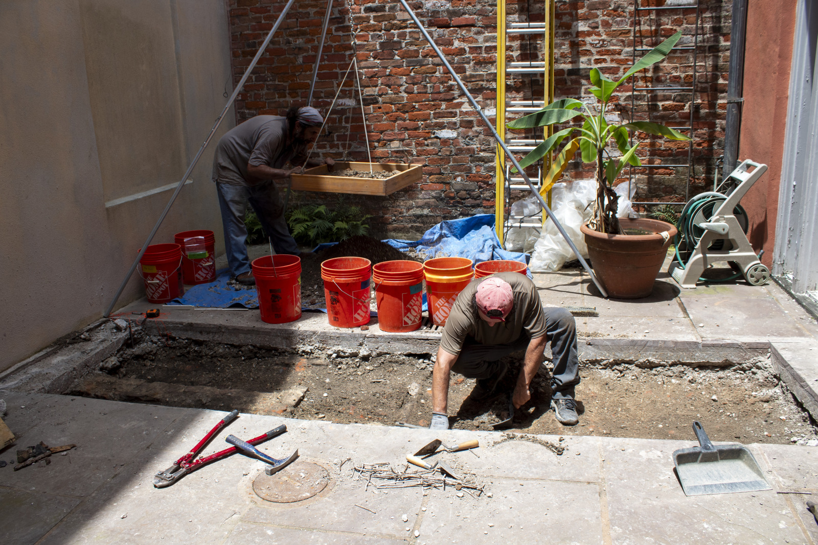 Archeologists dig in a French Quarter courtyard. A piece of concrete has been removed, exposing dirt below. Equipment and buckets are scattered around.