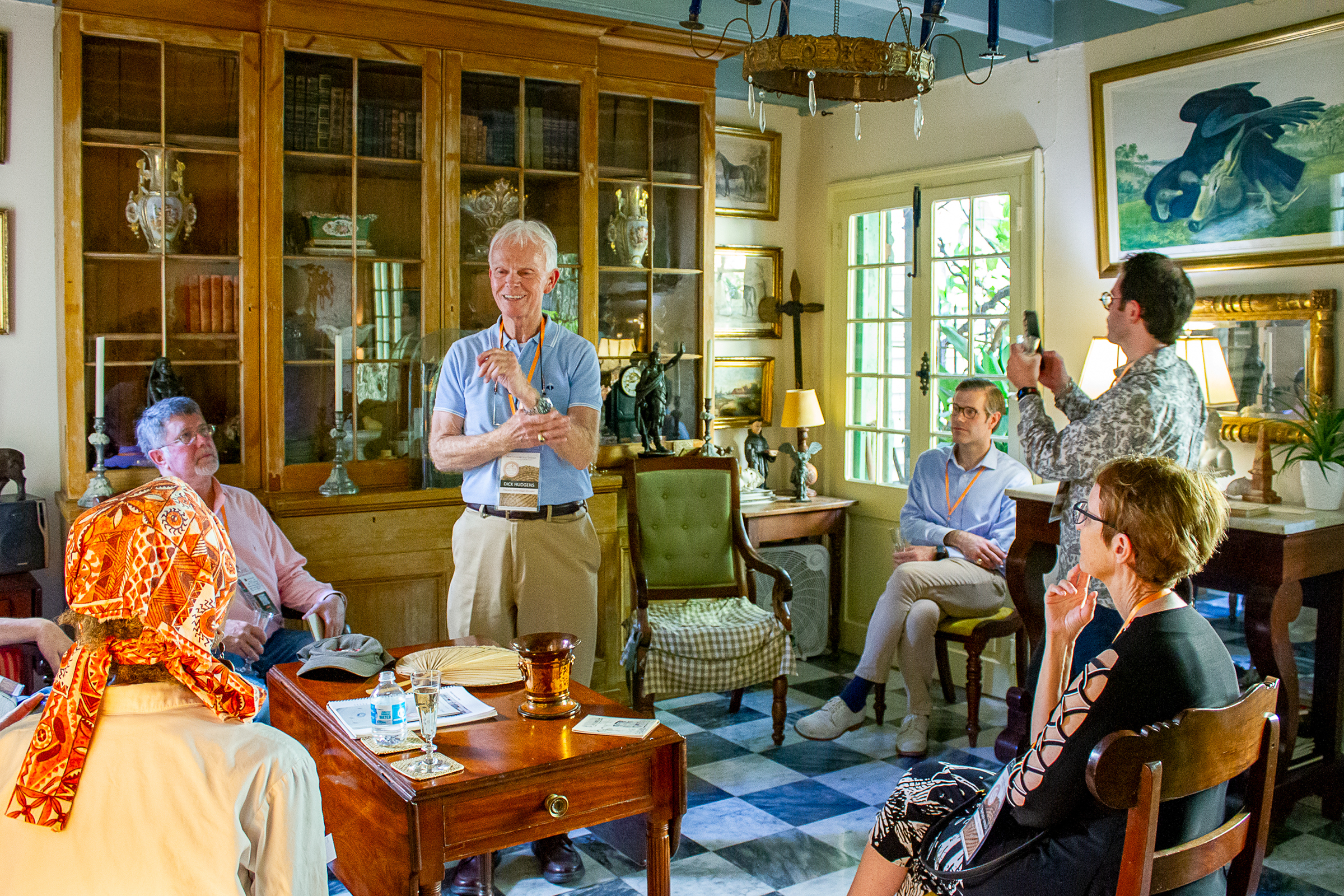 A group of people sit and stand in a cozy room filled with antique furniture and books. A man stands in the center, speaking animatedly. The room is warmly lit, with paintings and various decor items displayed on the walls and shelves.
