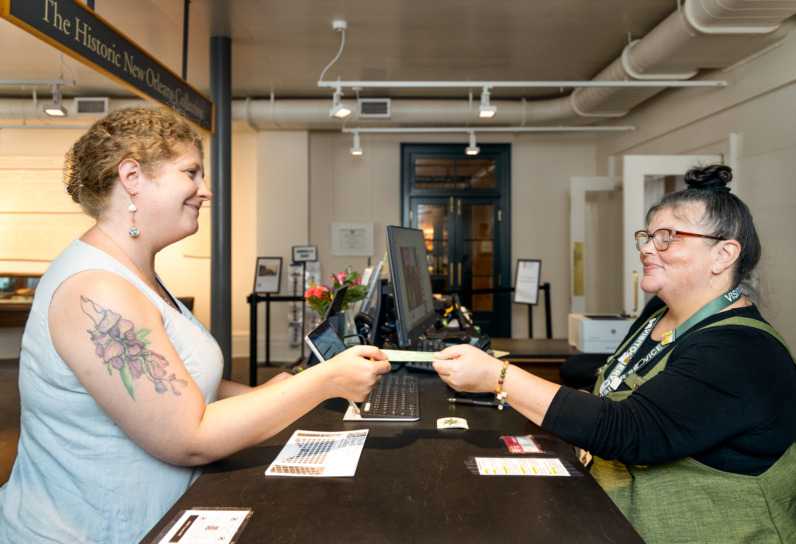 A woman with a flower tattoo on her arm stands at a reception desk, handing a card to a seated woman with glasses. They are smiling at each other in a warmly lit interior space. A sign above reads The Historic New Orl.