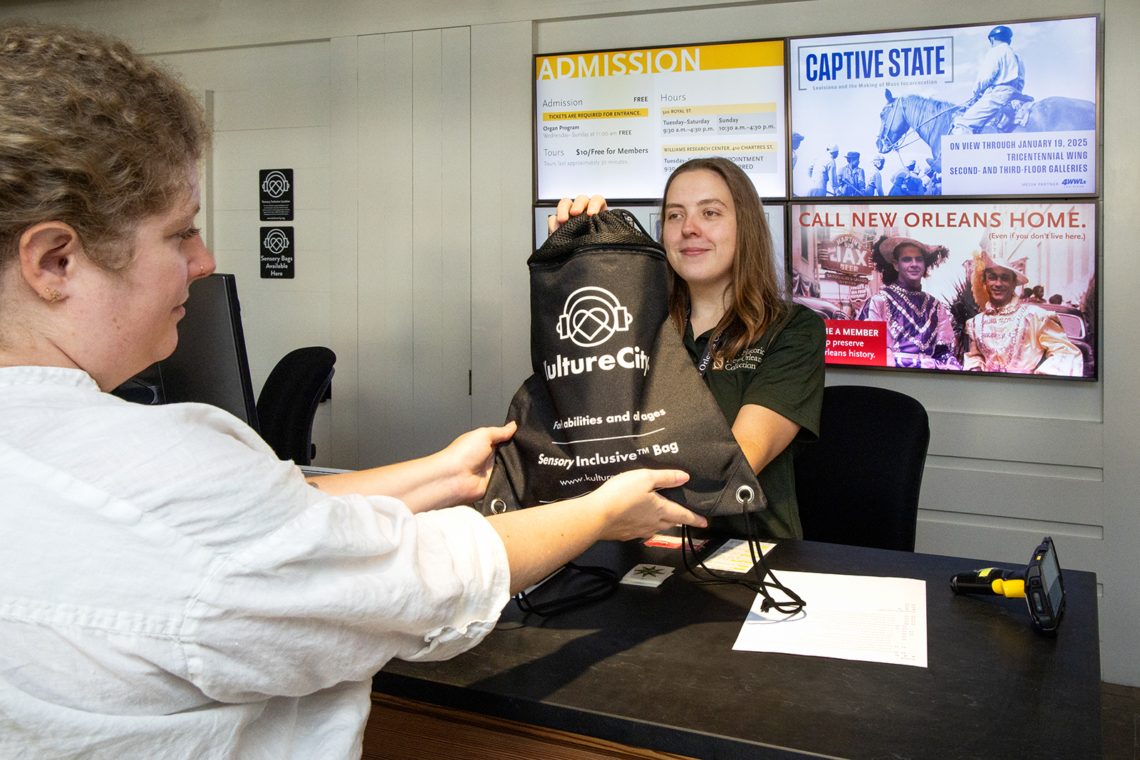 A woman at a reception desk hands a sensory inclusive bag labeled Kulture City to another woman. Behind them is an admission sign and a digital display with event posters.