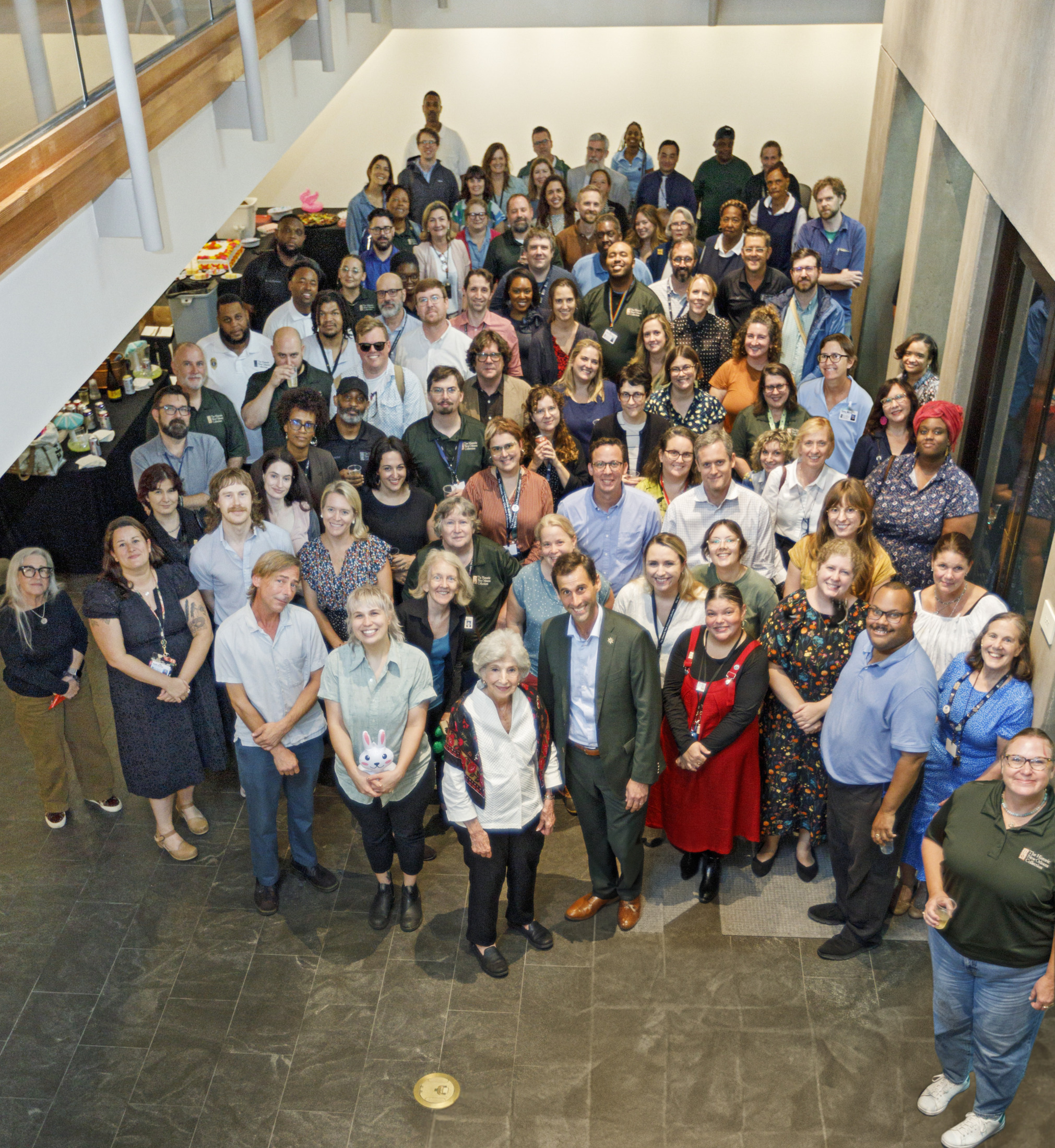 A large group of people of varying ages and attire gather indoors for a group photo. They are arranged in several rows, smiling and looking at the camera. The setting appears to be an event or meeting in a spacious area with a high ceiling.