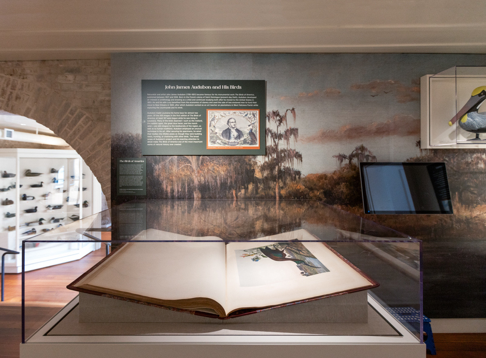 An open book displayed in a glass case within a museum exhibit. The background features a wall with a painted landscape and informational panels. A digital screen is visible to the side. Shelves with items are seen in another room through an archway.