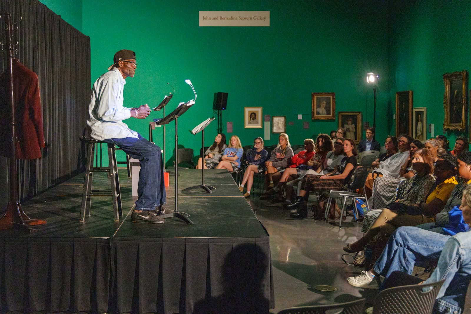 A man wearing a cap and glasses sits on a stool, reading from a script at a lectern, amid a theater setting with a backdrop of artworks. An audience sits attentively in a dimly lit, green-walled gallery.