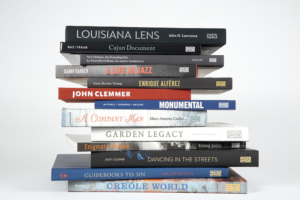 A stack of books with titles like Louisiana Lens, A Life in Jazz, Monumental, and Creole World. The books cover subjects related to Louisianas culture, art, and history. The spines are neatly aligned against a plain, white background.