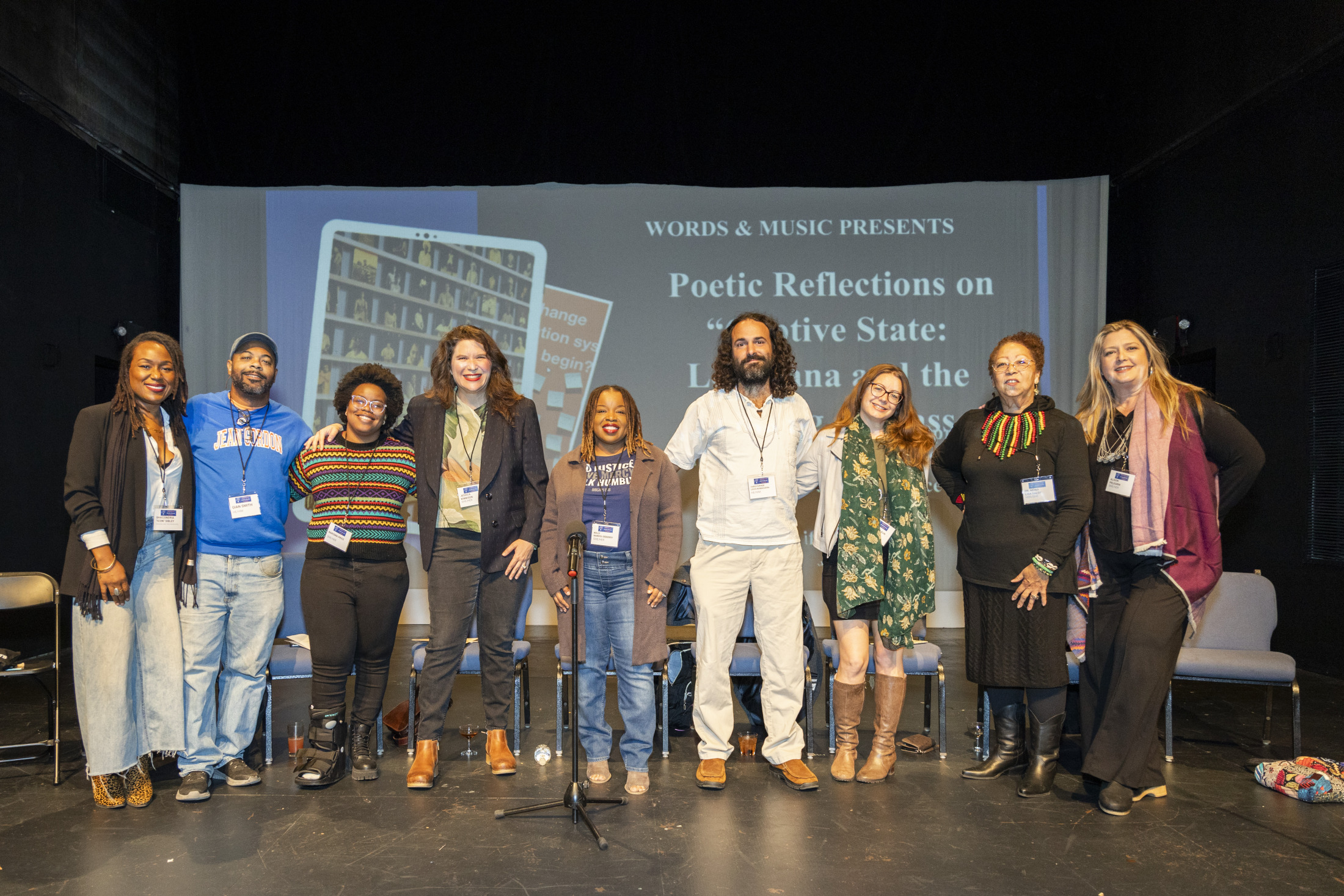 A diverse group of nine people standing on a stage, smiling at the camera. They are participating in an event titled Poetic Reflections on a Positive State: Finding Carolina In the... A screen behind them displays part of the event title.