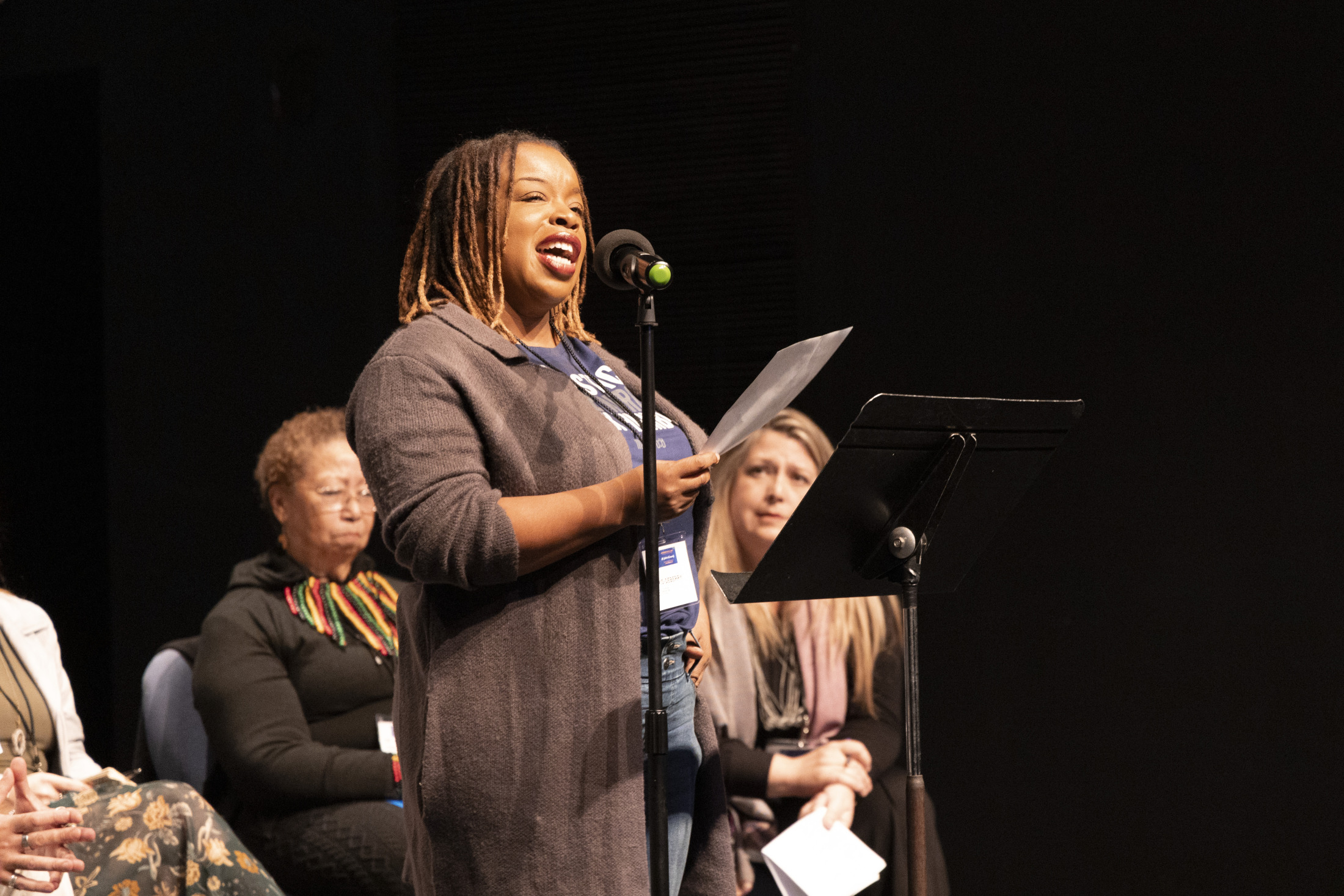 A speaker stands at a microphone holding a piece of paper, addressing an audience. She is in front of a group of seated individuals on stage. The setting appears to be a formal event or panel discussion.