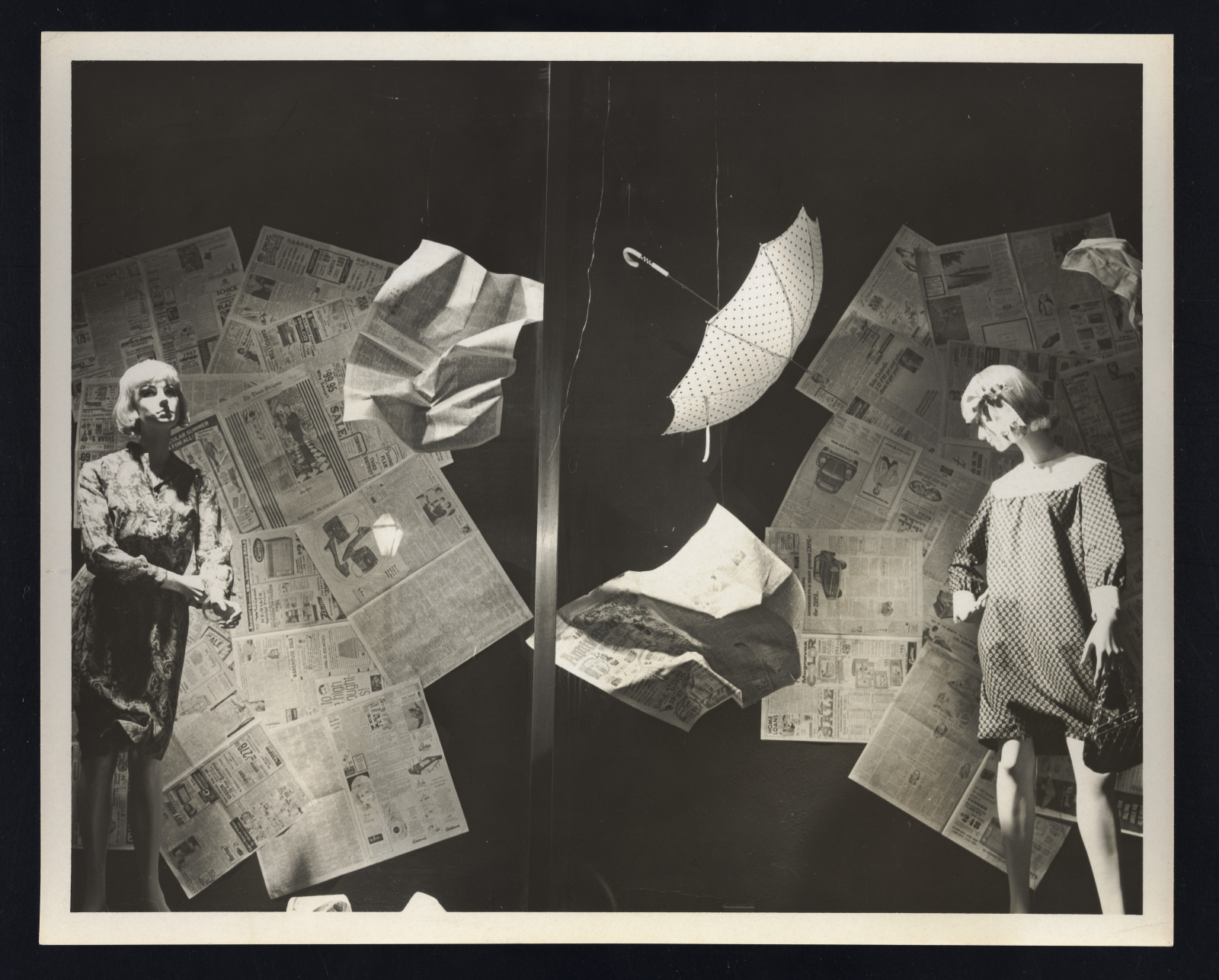 View of window display at Maison Blanche department store. Shows two mannequins in long-sleeved dresses, crumpled newspapers, and an umbrella suspended from the ceiling as if it were blowing in the wind.