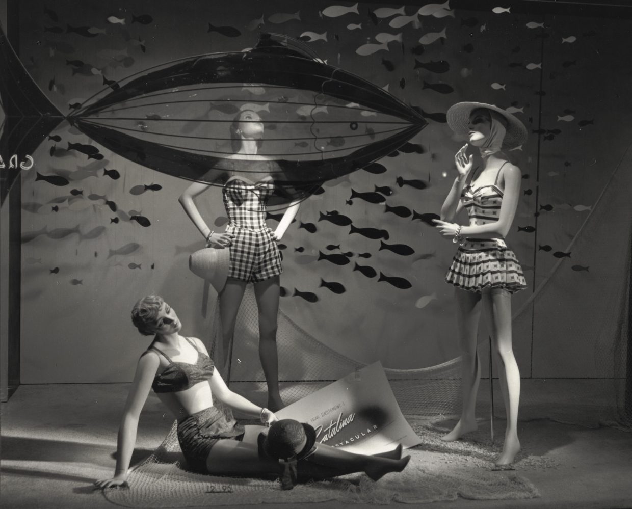 View of window display at Maison Blanche department store. The mannequins are modeling swimwear, including a two-piece. A school of fish mounted on the wall behind.