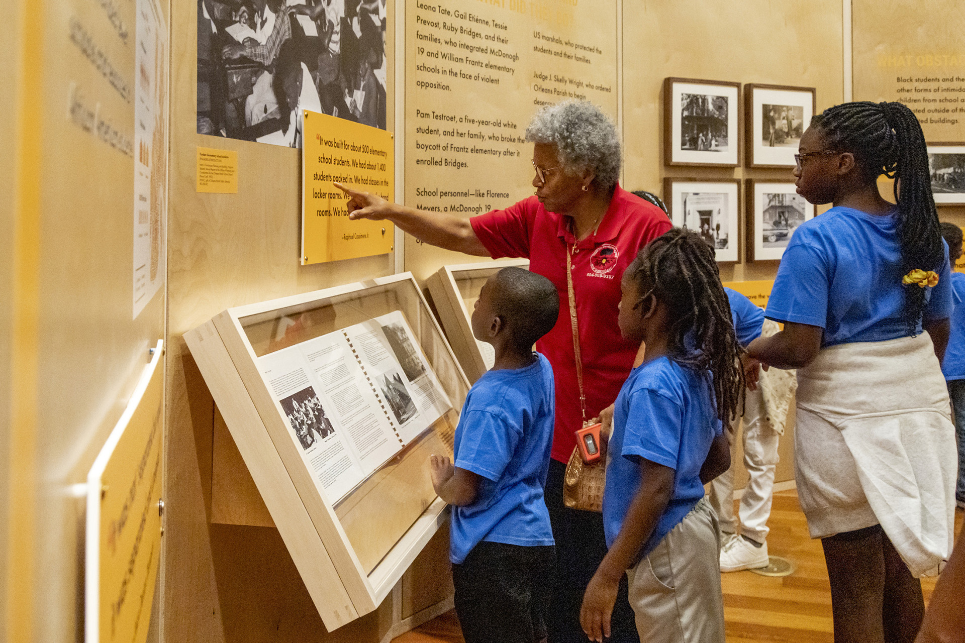A woman points at an installation at HNOC's "The Trail They Blazed" exhibition. Children are gathered around her.