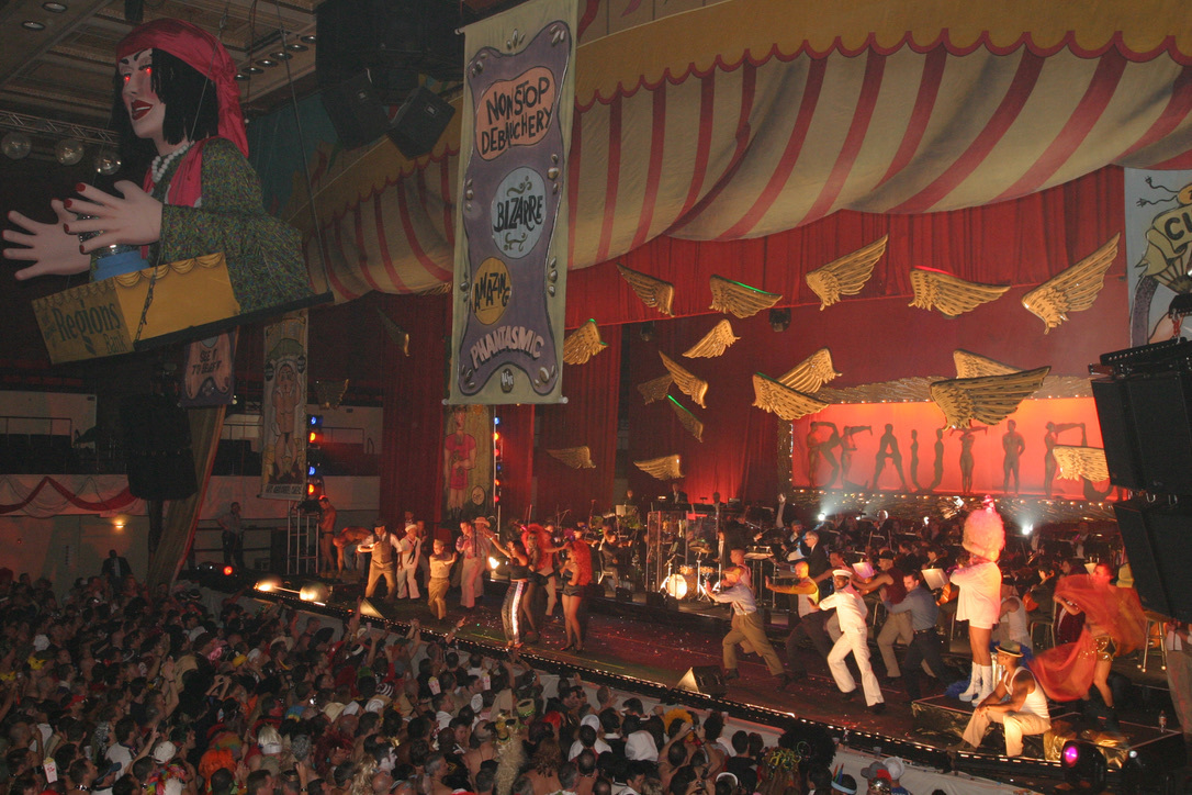 Photo of Halloween New Orleans (unknown year). A large stage is filled with costumed dancers with colorful banners hanging from the ceiling. A large crowd is enjoying the festivities.