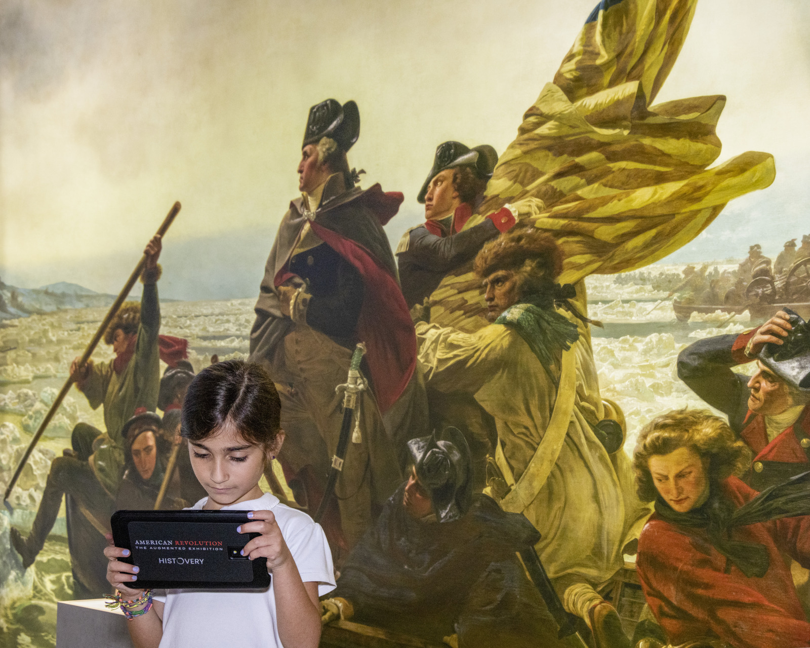 A young girl holds a Histopad tablet while standing in front of a backdrop of the famous painting of Washington crossing the Delaware River at "American Revolution: The Augmented Exhibition."