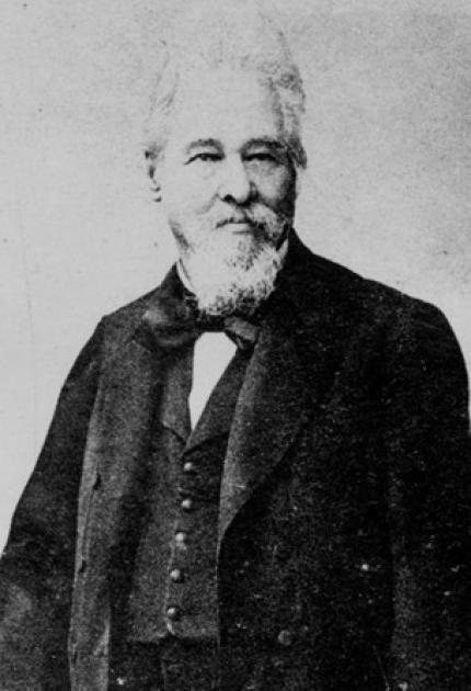 An older man with a white beard and hair, wearing a dark, formal suit with a bow tie, stands facing forward. The style suggests a historical portrait.