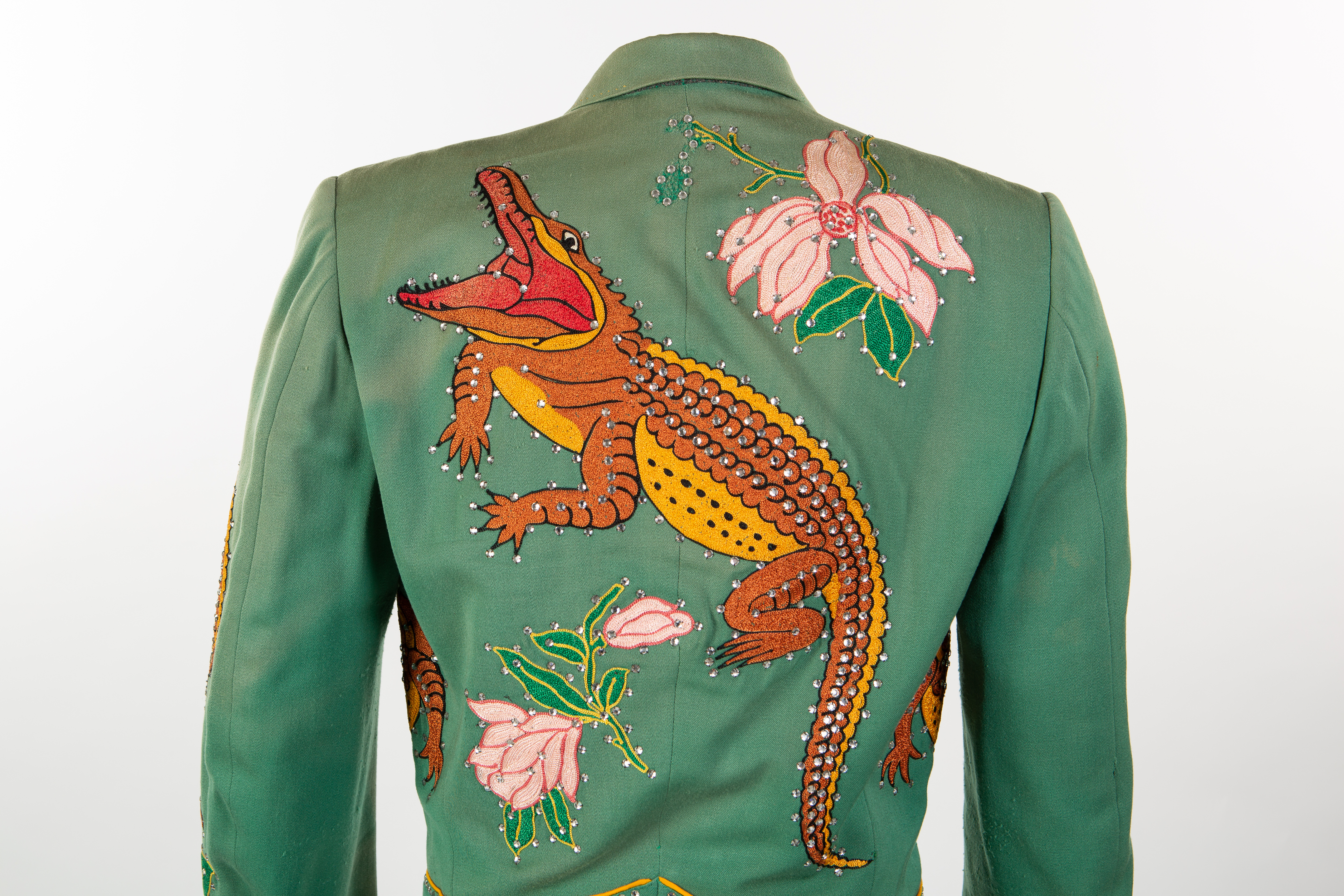 A green jacket featuring a colorful embroidered crocodile and pink flowers on the back. The detailed artwork includes beaded embellishments.