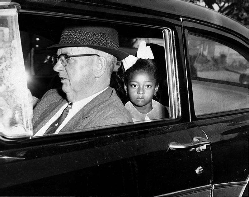 A young girl with a bow in her hair sits in the backseat of a car next to an older man wearing a hat and glasses. They both look out the window. The interior of the car is visible, with the exterior partially seen outside.