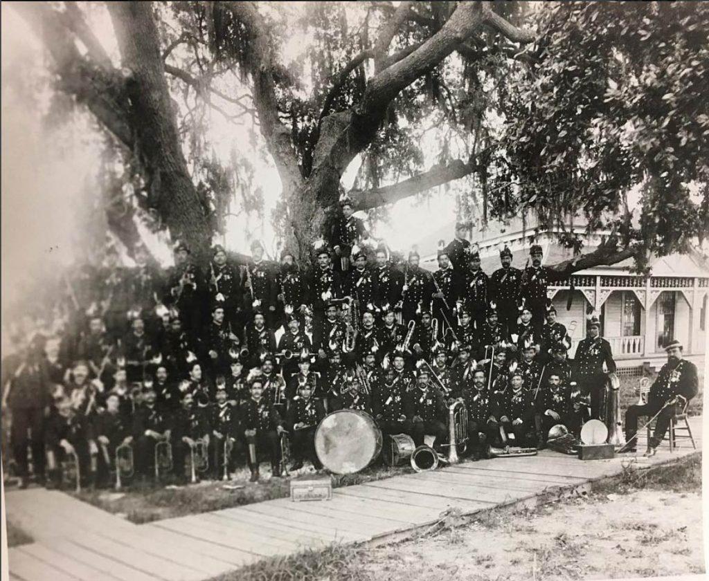 A large historical band poses with musical instruments under a large tree. The members are dressed in dark uniforms, and various instruments like drums and brass are visible. A building with a porch is in the background.