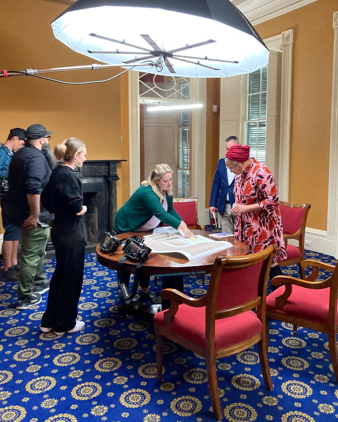 A group of people gathers around a table in a room with golden walls and blue patterned carpet. They appear to be discussing something on the table. A large light fixture is set up above them. The room is decorated with red chairs.