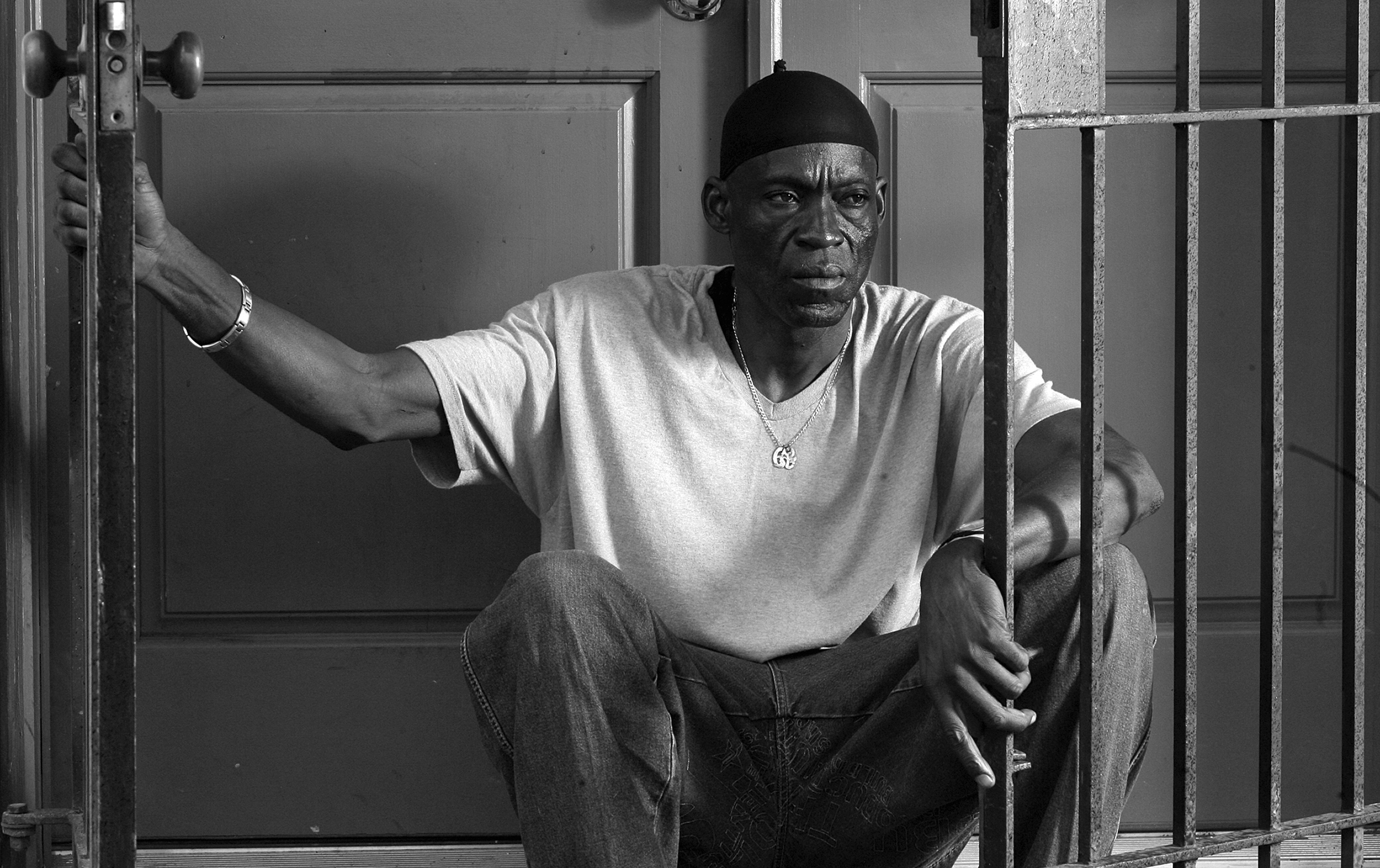 A black and white photo of a man sitting on a doorstep. He wears a light t-shirt, a do-rag, and a chain necklace. He is holding onto an iron gate with one hand and resting the other on his knee, looking pensively into the distance.