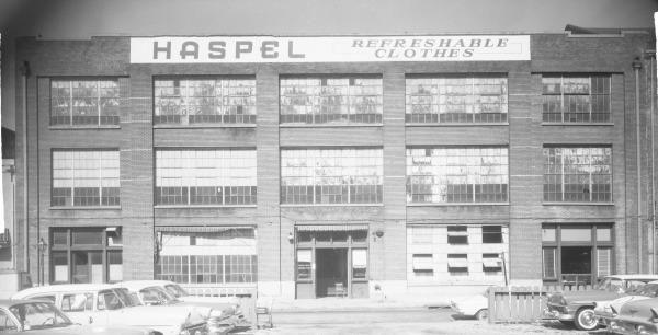 Black and white photo of a large brick building labeled HASPEL REFRESHABLE CLOTHES. The building has multiple rows of windows. Several vintage cars are parked in front.