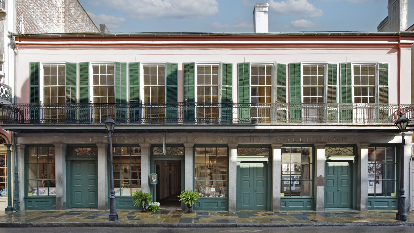A two-story building with a pink facade and green shutters in a historic style. The ground floor has large windows and doors surrounded by columns. There are potted plants at the entrance, and a balcony runs along the second floor.