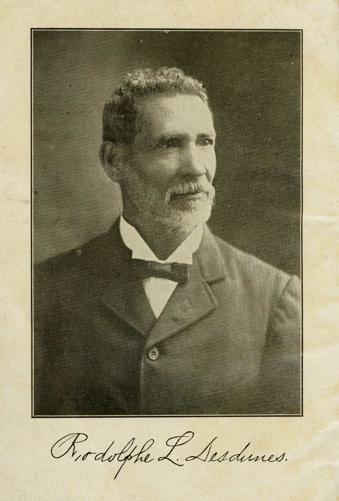 A sepia-toned portrait of an older man with short, curly hair and a mustache. He is wearing a dark suit with a bow tie. Below the photo is a handwritten signature in cursive.