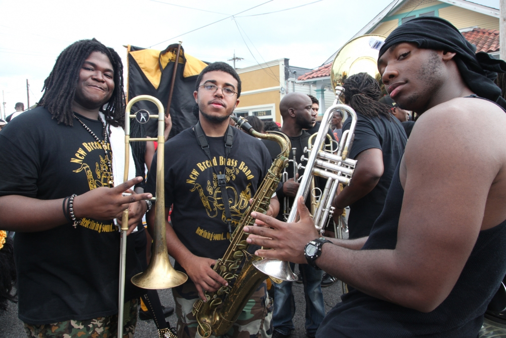 A group of young musicians wearing matching black shirts pose with their brass instruments on a street. They are part of a marching band, and some are holding trombones and a tuba. Buildings and a cloudy sky are visible in the background.