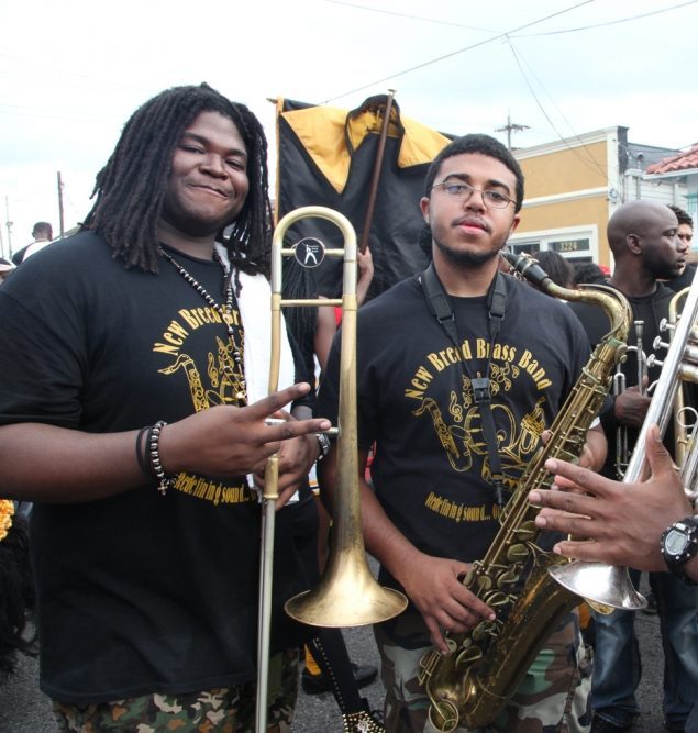 Two musicians from the New Breed Brass Band stand with their instruments at a street event. One holds a trombone and flashes a peace sign, while the other holds a saxophone. They wear band T-shirts and camouflage pants. A crowd is visible in the background.